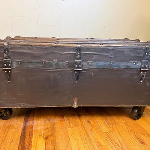 Puede incluir: Un ba&uacute;l antiguo con exterior gris oscuro, detalles met&aacute;licos y protectores de esquina. El ba&uacute;l tiene forma rectangular, se apoya sobre peque&ntilde;as ruedas y presenta bisagras y cierres met&aacute;licos. El marco de madera es visible en la parte superior y los laterales.