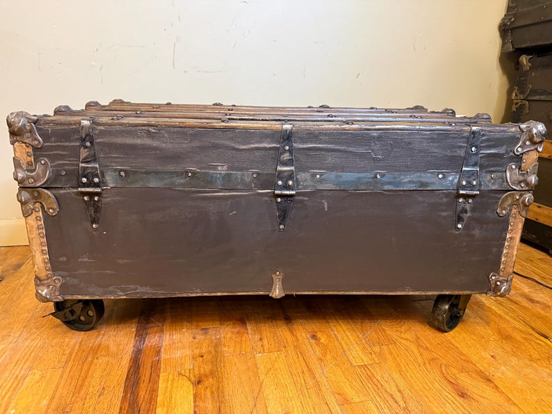 Puede incluir: Un ba&uacute;l antiguo con exterior gris oscuro, detalles met&aacute;licos y protectores de esquina. El ba&uacute;l tiene forma rectangular, se apoya sobre peque&ntilde;as ruedas y presenta bisagras y cierres met&aacute;licos. El marco de madera es visible en la parte superior y los laterales.