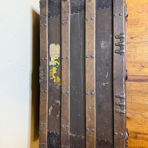 May include: Antique wooden trunk with metal accents. The trunk features dark wood panels, metal bands, and decorative corner pieces. The trunk has a weathered appearance with visible wear and tear, and a faded sticker. The trunk is a vintage storage item.
