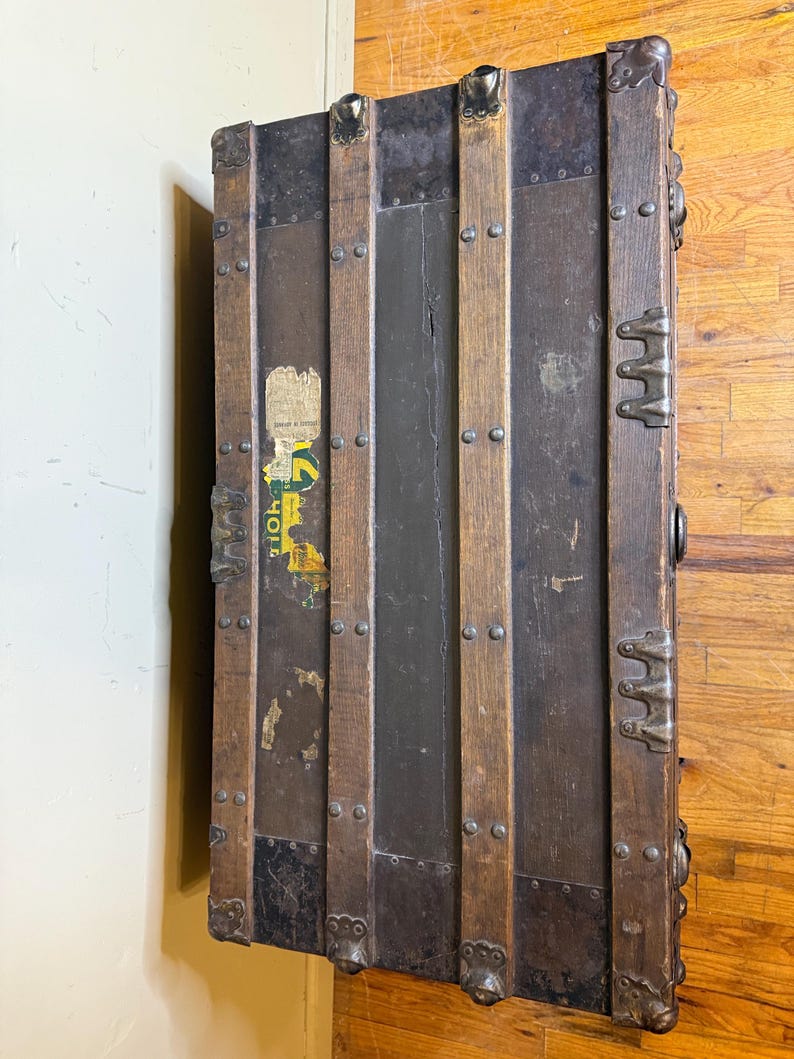 May include: Antique wooden trunk with metal accents. The trunk features dark wood panels, metal bands, and decorative corner pieces. The trunk has a weathered appearance with visible wear and tear, and a faded sticker. The trunk is a vintage storage item.