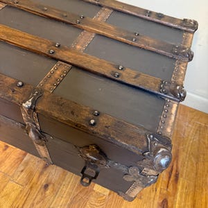 Puede incluir: Un ba&uacute;l antiguo con cuerpo gris oscuro y listones de madera. El ba&uacute;l presenta protectores de esquina met&aacute;licos y un pestillo. La madera tiene un acabado natural y los detalles met&aacute;licos est&aacute;n envejecidos, d&aacute;ndole un aspecto vintage.