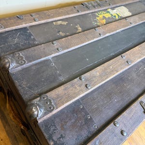 May include: Antique wooden trunk with metal accents. The trunk features dark wood slats, metal corner protectors, and a distressed finish. The metal is dark gray, and the wood is a medium brown. The trunk has a vintage aesthetic.
