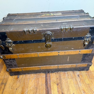 May include: An antique wooden trunk with metal hardware. The trunk is primarily brown with black accents on the corners and a black stripe. The trunk has metal latches and a lock. The trunk is rectangular and sits on a wooden floor.