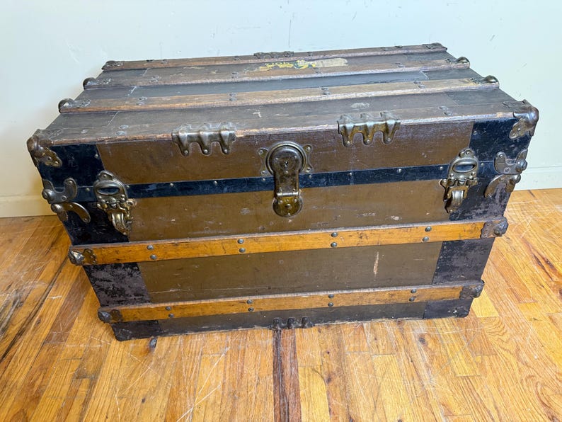 May include: An antique wooden trunk with metal hardware. The trunk is primarily brown with black accents on the corners and a black stripe. The trunk has metal latches and a lock. The trunk is rectangular and sits on a wooden floor.