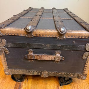 Puede incluir: Un ba&uacute;l negro antiguo con listones de madera y detalles met&aacute;licos. El ba&uacute;l tiene un asa de cuero y peque&ntilde;as ruedas. Las esquinas y los bordes est&aacute;n reforzados con metal. Un art&iacute;culo de almacenamiento vintage.