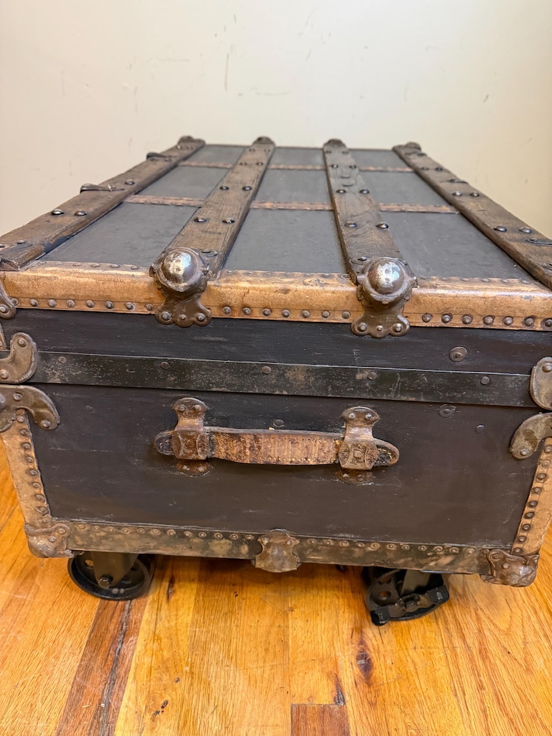 Puede incluir: Un ba&uacute;l negro antiguo con listones de madera y detalles met&aacute;licos. El ba&uacute;l tiene un asa de cuero y peque&ntilde;as ruedas. Las esquinas y los bordes est&aacute;n reforzados con metal. Un art&iacute;culo de almacenamiento vintage.