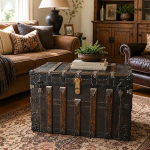 May include: An antique black trunk with metal accents and wooden straps serves as a coffee table. The trunk is in a living room with a brown sofa, patterned pillows, and a patterned rug. Books and a plant sit on top of the trunk.