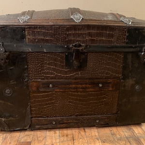 Pode incluir: Um ba&uacute; vintage de couro marrom com detalhes e ferragens met&aacute;licas. O ba&uacute; tem uma tampa abobadada e &eacute; seguro com fechos de metal.