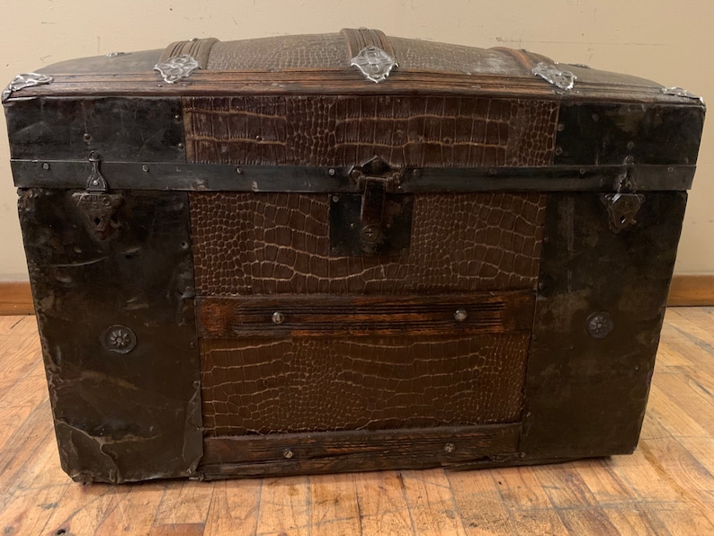 Pode incluir: Um ba&uacute; vintage de couro marrom com detalhes e ferragens met&aacute;licas. O ba&uacute; tem uma tampa abobadada e &eacute; seguro com fechos de metal.