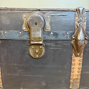 Puede incluir: Primer plano de un ba&uacute;l antiguo con exterior gris oscuro. El ba&uacute;l presenta un gran cierre met&aacute;lico ornamentado y detalles met&aacute;licos decorativos. Las correas de cuero con tachuelas met&aacute;licas se suman a la est&eacute;tica vintage. El ba&uacute;l est&aacute; sobre una superficie de madera.