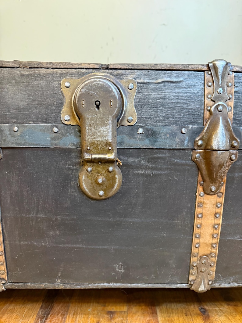 Puede incluir: Primer plano de un ba&uacute;l antiguo con exterior gris oscuro. El ba&uacute;l presenta un gran cierre met&aacute;lico ornamentado y detalles met&aacute;licos decorativos. Las correas de cuero con tachuelas met&aacute;licas se suman a la est&eacute;tica vintage. El ba&uacute;l est&aacute; sobre una superficie de madera.