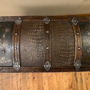 Pode incluir: Um ba&uacute; de madeira de estilo vintage com acabamento em couro marrom e detalhes em metal. O ba&uacute; tem uma parte superior curva e &eacute; decorado com desenhos florais de metal.