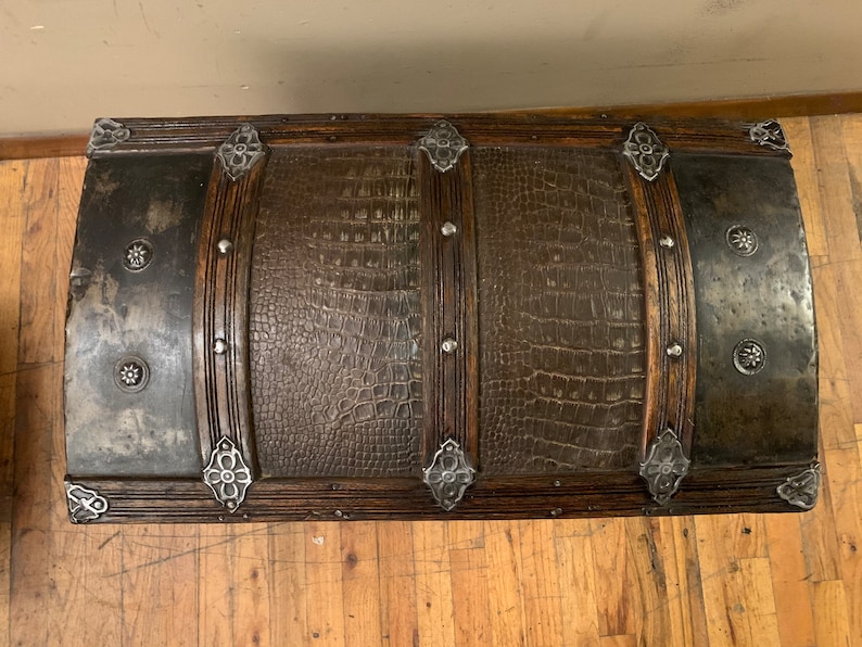 Pode incluir: Um ba&uacute; de madeira de estilo vintage com acabamento em couro marrom e detalhes em metal. O ba&uacute; tem uma parte superior curva e &eacute; decorado com desenhos florais de metal.