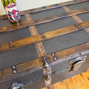 Puede incluir: Ba&uacute;l de madera antiguo con herrajes met&aacute;licos y correas de cuero. El ba&uacute;l tiene un exterior gris oscuro con detalles de madera y remaches met&aacute;licos. Una botella verde con una etiqueta descansa encima.