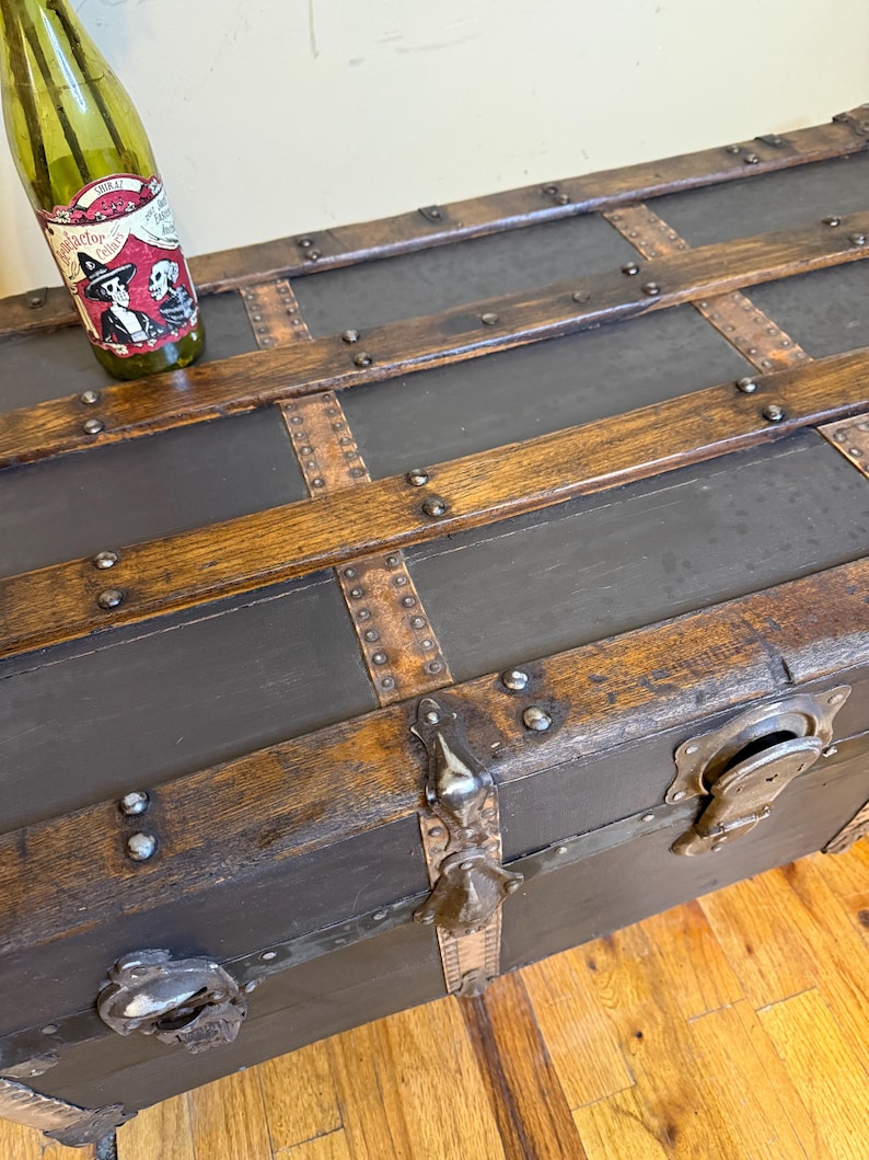 Puede incluir: Ba&uacute;l de madera antiguo con herrajes met&aacute;licos y correas de cuero. El ba&uacute;l tiene un exterior gris oscuro con detalles de madera y remaches met&aacute;licos. Una botella verde con una etiqueta descansa encima.