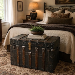 May include: An antique black trunk with metal accents and leather straps sits at the foot of a bed. The trunk is topped with books and a potted plant. The bedroom has a neutral color palette with a patterned rug.
