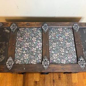 May include: A rectangular wooden storage trunk featuring dark wood slats, black side panels with a distressed finish, and decorative silver-toned metal corner accents. The top surface includes two panels covered in a dark floral fabric pattern.