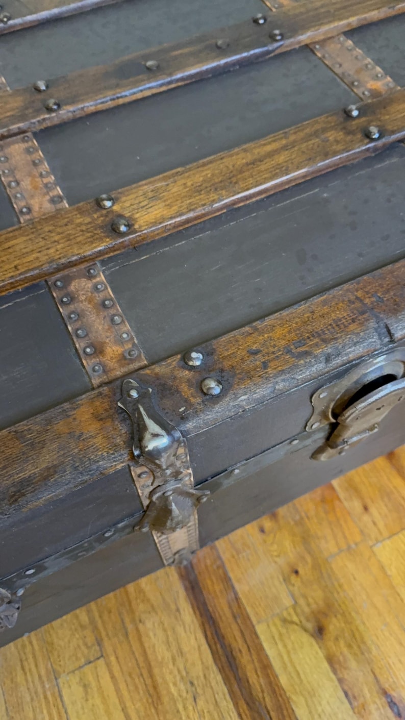 Puede incluir: Ba&uacute;l antiguo con paneles gris oscuro y molduras de madera. El ba&uacute;l presenta detalles met&aacute;licos, incluyendo protectores de esquina y remaches. La moldura de madera tiene un aspecto c&aacute;lido y envejecido.
