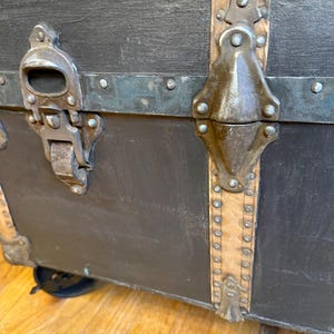 Puede incluir: Primer plano de un ba&uacute;l negro vintage con herrajes met&aacute;licos. El ba&uacute;l presenta protectores de esquina met&aacute;licos, un pestillo met&aacute;lico y correas de cuero. El ba&uacute;l est&aacute; sobre ruedas y tiene un marco de madera.