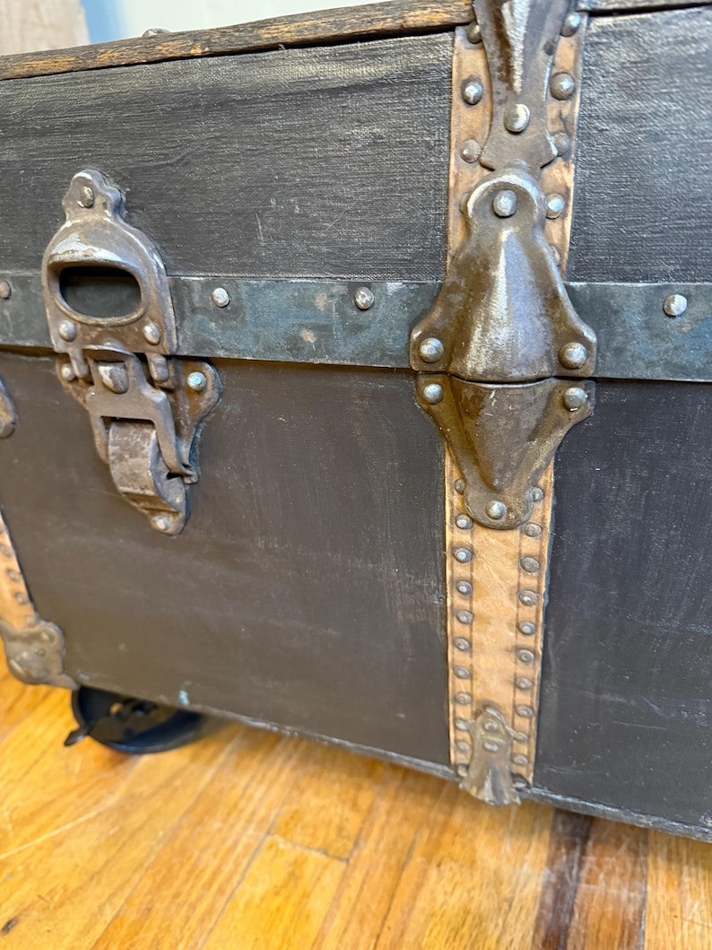 Puede incluir: Primer plano de un ba&uacute;l negro vintage con herrajes met&aacute;licos. El ba&uacute;l presenta protectores de esquina met&aacute;licos, un pestillo met&aacute;lico y correas de cuero. El ba&uacute;l est&aacute; sobre ruedas y tiene un marco de madera.