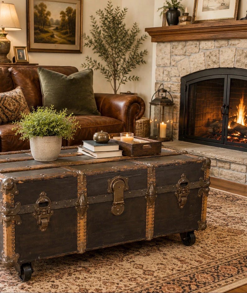 Puede incluir: Un ba&uacute;l vintage marr&oacute;n oscuro con detalles met&aacute;licos sirve como mesa de centro en un sal&oacute;n acogedor. El ba&uacute;l tiene cierres met&aacute;licos decorativos y est&aacute; sobre ruedas. Un sof&aacute; de cuero, una chimenea y una alfombra completan el espacio c&aacute;lido.