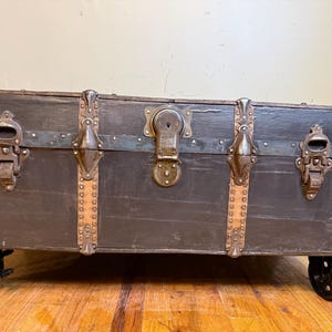 Puede incluir: Un ba&uacute;l de madera antiguo con acabado oscuro, detalles met&aacute;licos y protectores de esquina. El ba&uacute;l presenta una cerradura central, pestillos met&aacute;licos y est&aacute; montado sobre peque&ntilde;as ruedas negras. El exterior del ba&uacute;l es de color gris oscuro.