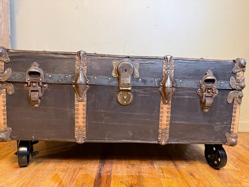 Puede incluir: Un ba&uacute;l de madera antiguo con acabado oscuro, detalles met&aacute;licos y protectores de esquina. El ba&uacute;l presenta una cerradura central, pestillos met&aacute;licos y est&aacute; montado sobre peque&ntilde;as ruedas negras. El exterior del ba&uacute;l es de color gris oscuro.