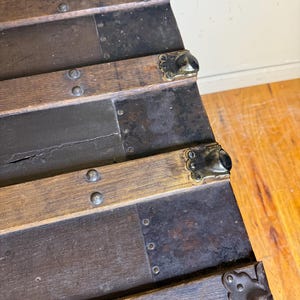 May include: Close-up of an antique wooden trunk with metal accents. The trunk features dark brown wooden slats, reinforced with metal strips and decorative corner pieces. Rivets secure the metal and wood components, showcasing a vintage aesthetic.