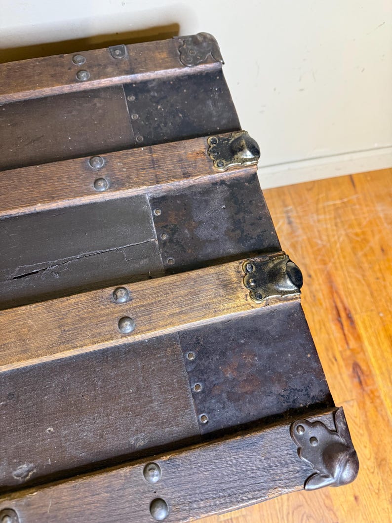 May include: Close-up of an antique wooden trunk with metal accents. The trunk features dark brown wooden slats, reinforced with metal strips and decorative corner pieces. Rivets secure the metal and wood components, showcasing a vintage aesthetic.