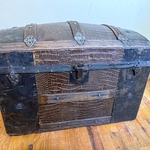 May include: A vintage dome-top trunk featuring a dark brown crocodile-textured leather finish, reinforced with black metal bands, decorative silver-toned metal corner accents, and a central metal latch mechanism on the front panel.