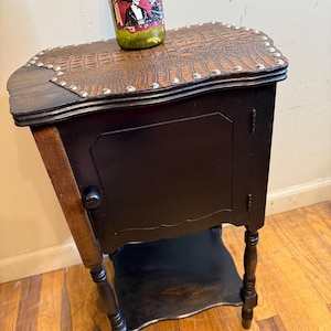 May include: A small, black wooden cabinet with a brown, textured top and silver studs. The cabinet has a single door and a lower shelf. A green bottle with a label featuring a skeleton and a man in a hat is on top.