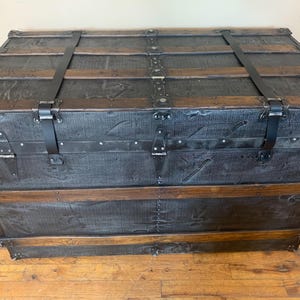 K&ouml;nnte beinhalten: Ein Vintage-Koffer aus schwarzem Leder mit Holzakzenten und Metallbeschl&auml;gen. Der Koffer hat zwei Riemen und ist geschlossen.