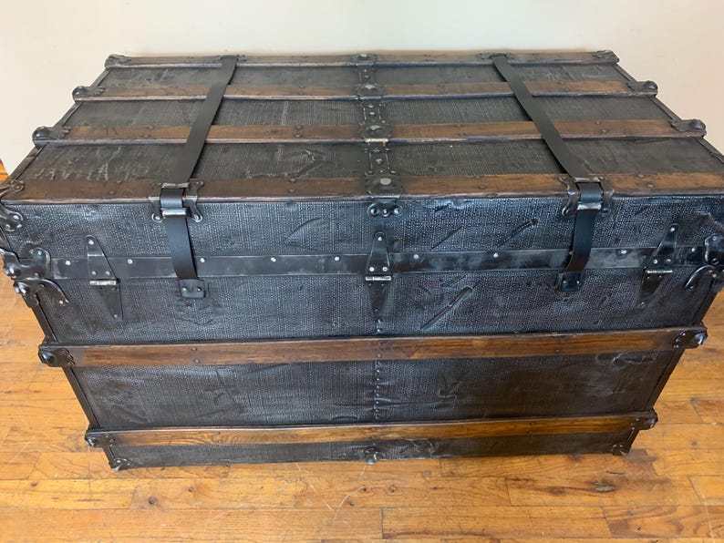 K&ouml;nnte beinhalten: Ein Vintage-Koffer aus schwarzem Leder mit Holzakzenten und Metallbeschl&auml;gen. Der Koffer hat zwei Riemen und ist geschlossen.