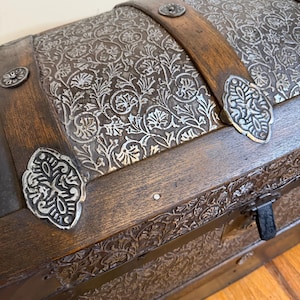 May include: A wooden storage chest with a domed lid featuring dark brown wood slats and silver-toned metal panels embossed with intricate floral patterns. The chest includes decorative silver metal corner accents and a black metal latch mechanism.