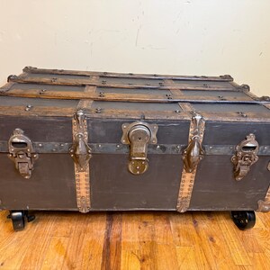 Puede incluir: Un ba&uacute;l antiguo con exterior gris oscuro, listones de madera y herrajes met&aacute;licos. El ba&uacute;l presenta una gran cerradura central y cierres met&aacute;licos decorativos. Descansa sobre peque&ntilde;as ruedas negras, lo que facilita su traslado.
