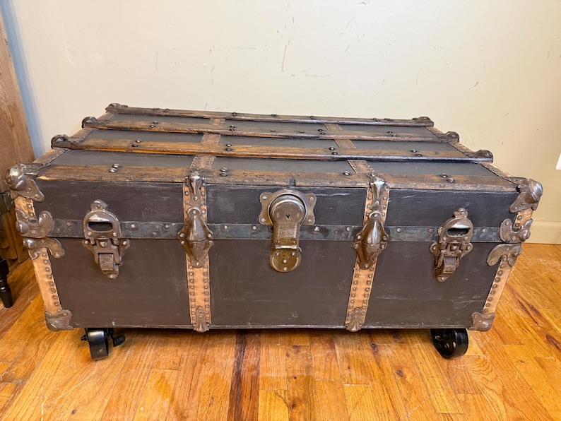 Puede incluir: Un ba&uacute;l antiguo con exterior gris oscuro, listones de madera y herrajes met&aacute;licos. El ba&uacute;l presenta una gran cerradura central y cierres met&aacute;licos decorativos. Descansa sobre peque&ntilde;as ruedas negras, lo que facilita su traslado.