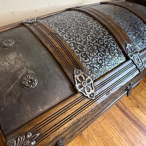 May include: A wooden storage chest with a domed lid featuring dark metal panels. The metal surfaces are decorated with embossed floral patterns and silver-toned metal corner accents, straps, and circular studs across the exterior.