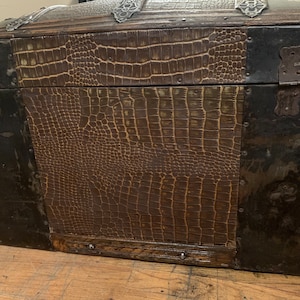 Pode incluir: Um ba&uacute; de madeira vintage com uma cobertura de couro marrom com estampa de crocodilo. O ba&uacute; tem detalhes e dobradi&ccedil;as de metal.