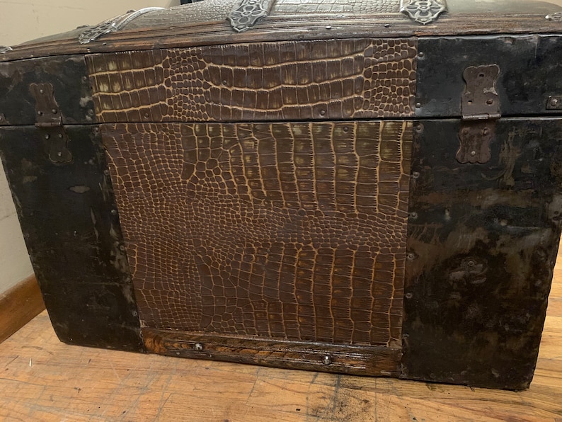 Pode incluir: Um ba&uacute; de madeira vintage com uma cobertura de couro marrom com estampa de crocodilo. O ba&uacute; tem detalhes e dobradi&ccedil;as de metal.