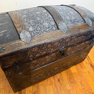 May include: A large, rectangular storage chest with a domed lid, featuring a dark brown and black finish with intricate floral patterns. The chest is reinforced with dark wood slats, silver-toned metal corner accents, and metal latches on the front.