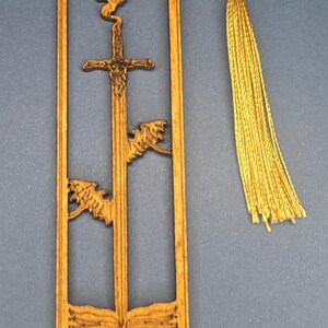 May include: A rectangular wooden bookmark featuring a laser-cut design of a sword piercing an open book, with two small dragon figures and the text "Knowledge Is Power" engraved at the bottom. A gold-colored cord tassel is attached to the top.