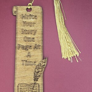 Puede incluir: Un marcapáginas de madera rectangular con forma de pergamino y una borla dorada. La madera está grabada con el texto "Write Your Story One Page At A Time" y una ilustración de un libro abierto con una pluma sobre las páginas.