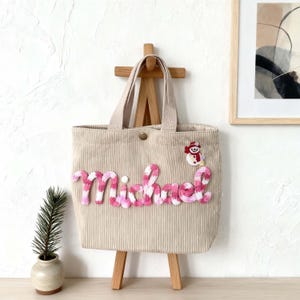 Può includere: Una borsa tote in velluto a coste beige con il nome Michael ricamato in filato sfumato rosa e bianco. Un piccolo pupazzo di neve ricamato con sciarpa e cappello rossi è posizionato sopra il nome. La borsa ha due manici e una chiusura a pressione.