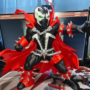 Pode incluir: Uma figura de ação do Spawn com um traje preto e branco, uma capa vermelha grande, uma fivela de cinto prateada em forma de caveira, correntes prateadas no peito e na cintura, e punhos vermelhos com espinhos nos pulsos e pernas.