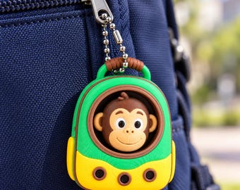 May include: A small, colorful keychain shaped like a backpack with a brown monkey face peeking out from a circular window. The keychain features a bright green top, a yellow bottom with three black dots, and a brown handle, attached by a silver chain.