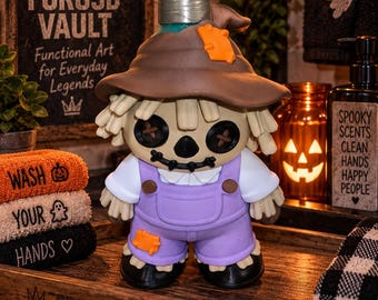 May include: A soap dispenser shaped like a scarecrow with a brown hat, a light purple pair of overalls, black button eyes, and a stitched mouth. The figure features a silver-toned pump mechanism on top and includes orange patches on the hat and leg.