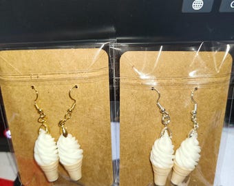 May include: Two pairs of dangle earrings shaped like white soft-serve ice cream cones. One pair features gold-toned metal hooks and the other pair features silver-toned metal hooks. Each pair is individually packaged on a brown cardboard display card.