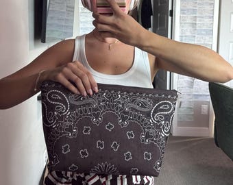 Könnte beinhalten: Eine dunkelbraune Reißverschlusstasche mit weißem Paisley-Bandana-Muster. Die rechteckige Tasche hat eine flache Oberkante mit Reißverschluss und einen breiteren Boden und zeigt ein traditionelles, komplexes Blumen- und Tropfenmuster.