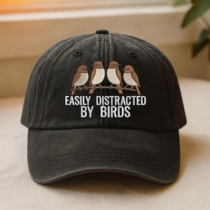 Könnte beinhalten: Eine schwarze Baseballkappe mit einer Stickerei von vier braun-weißen Vögeln, die auf einem Ast sitzen. Unter den Vögeln steht der Text EASILY DISTRACTED BY BIRDS in weißen Großbuchstaben. Die Kappe hat einen gebogenen Schirm und eine Krone.