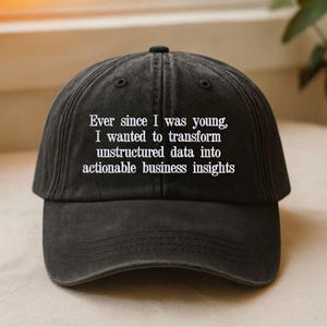 May include: A black baseball cap with a washed-out finish featuring white embroidered text that reads: Ever since I was young, I wanted to transform unstructured data into actionable business insights. The cap has a curved brim and six panels.
